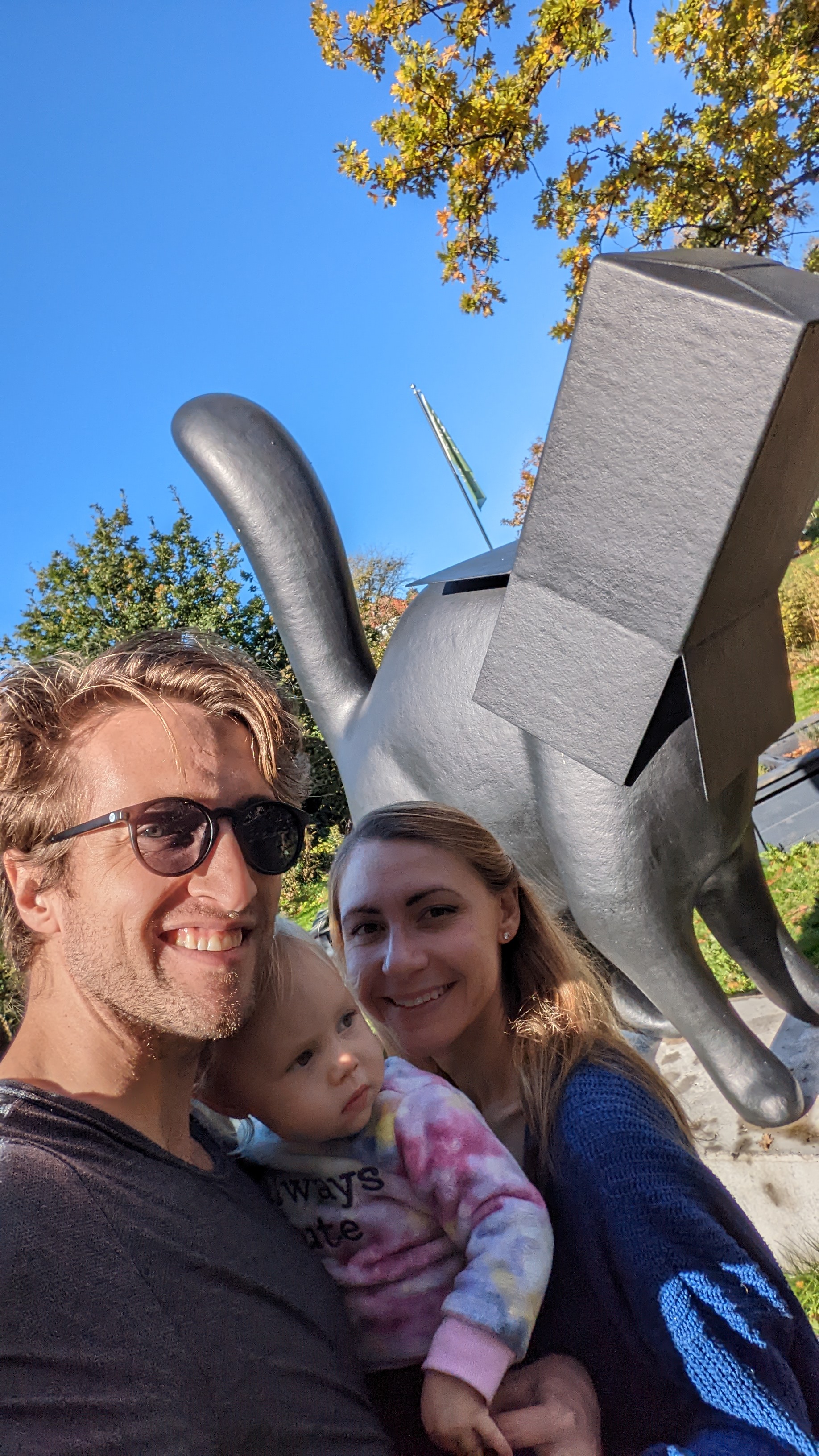 A man, woman and child smiling in front of a statue of a cat with a takeout box on it's head